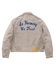 A beige canvas utility jacket shown from the back, featuring 'In Harmony We Trust' embroidered in blue script across the upper back and a small tan leather patch near the lower right hem.
