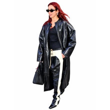 A woman modeling a black faux-leather longline coat over a black top and white pants with a black handbag, set against a plain white background.