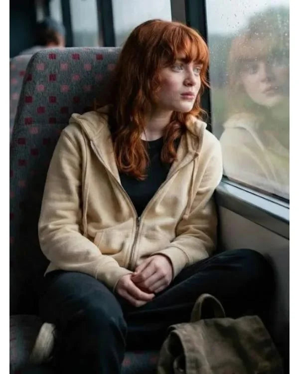 A person with red hair sits in a train seat by a window, wearing a beige zip-up hoodie over a black top and dark pants. A matching beige bag rests on the seat beside them.