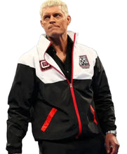 A man wearing a black and white zip-up jacket with red trim and WWE branding, standing in a ring environment.