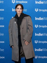A woman wearing a long taupe-gray double-breasted coat over a black outfit, standing in front of a blue Dropbox and IndieWire event backdrop.