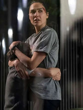 A woman in a gray t-shirt and dark pants holds a child close to her chest in a dim, nighttime setting.