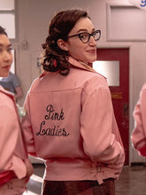 A woman wearing a pink jacket with 'Pink Ladies' embroidered on the back, standing in a school hallway.