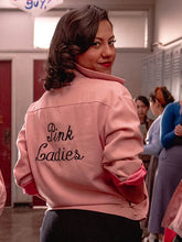 A woman in a pink jacket with 'Pink Ladies' embroidered on the back, standing in a school hallway with lockers in the background.