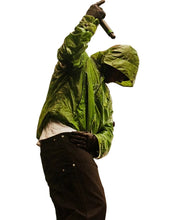 A person wearing a shiny olive-green hooded jacket, dark pants, and gloves, holding a microphone above their head against a plain light gray background.