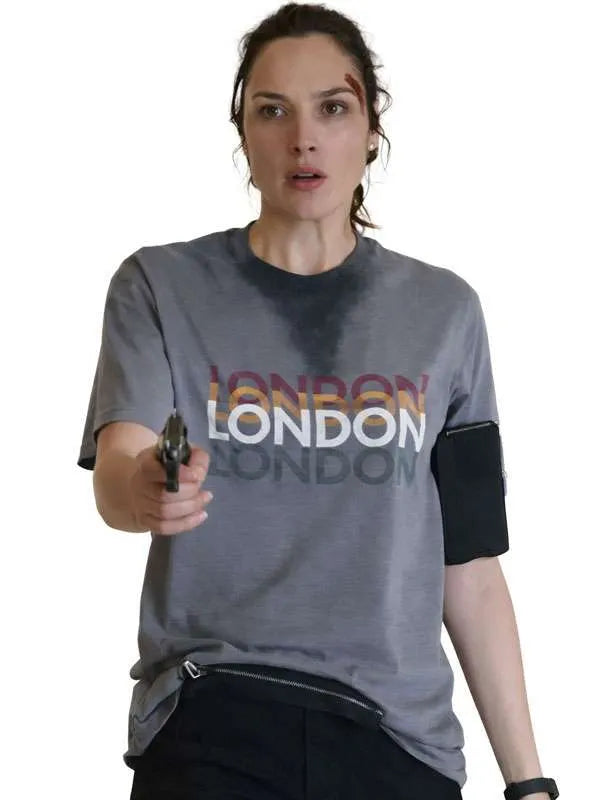A woman wearing a gray graphic T-shirt with 'LONDON' printed across the chest in a multicolor gradient and white text, paired with black pants. She is holding a gun and posed against a plain white background.