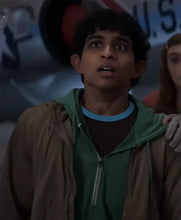 A young man with dark curly hair wearing a brown zip-up jacket over a green hoodie and a blue undershirt, looking surprised and upward. A woman is partially visible behind him.