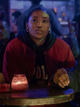A woman wearing a red and black hoodie sits at a table in a dimly lit bar or cafe, looking off-camera with a candle glowing on the table in front of her.