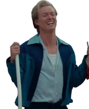 A man laughing and smiling while wearing a blue and teal color-blocked jacket over a light blue button-up shirt, holding onto white poles in front of a white wire mesh gate.