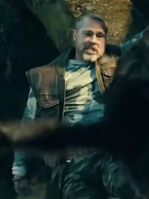 A man with a gray beard wearing a brown leather vest over a blue-gray shirt, standing in a dark, rocky outdoor setting.