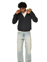 A man wearing a black zip-up hoodie with a beige faux-fur trimmed hood, light-wash blue jeans, and a white t-shirt, standing against a plain off-white studio background.