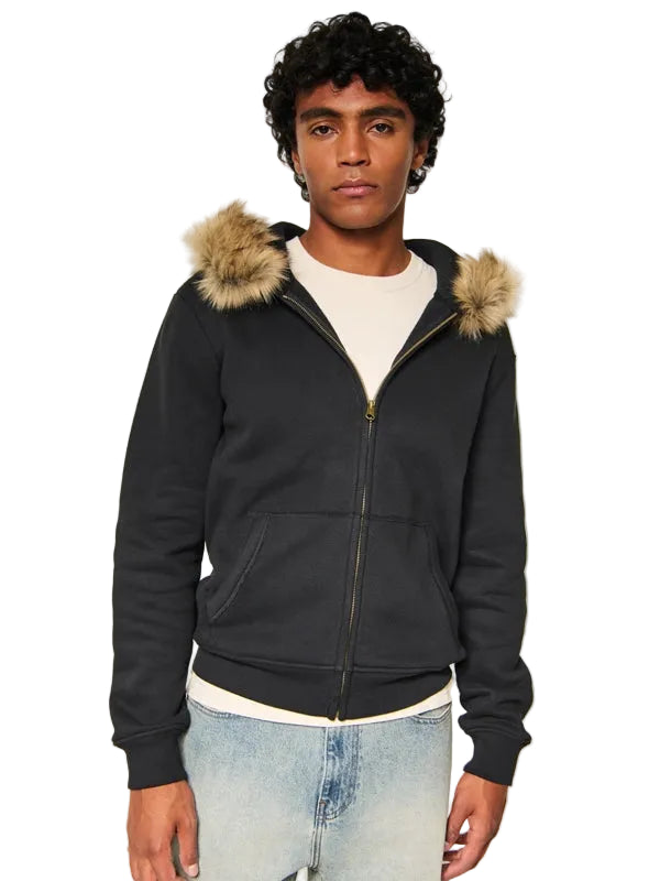 A man wearing a black zip-up hoodie with a tan faux-fur trimmed hood, layered over a white t-shirt and light-wash blue jeans, photographed against a neutral light gray background.