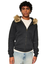 A man wearing a black zip-up hoodie with a tan faux-fur trimmed hood, layered over a white t-shirt and light-wash blue jeans, photographed against a neutral light gray background.