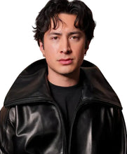 A man with dark hair wearing a black leather jacket over a black t-shirt, photographed against a textured, warm-toned brick wall background.