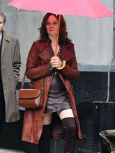 A woman walking under a pink umbrella wearing a long brown leather coat, a dark gray mini skirt, black over-the-knee boots, and carrying a brown leather shoulder bag.
