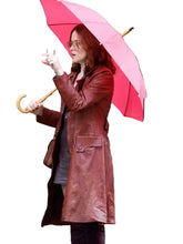 A woman standing on a city street holding a bright pink umbrella and wearing a long reddish-brown leather coat over a dark outfit, with a shop window and black-and-white pole in the background.