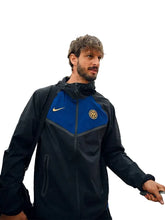 A man wearing a black and blue Inter Milan athletic jacket with a Nike swoosh, standing in front of an airplane door.