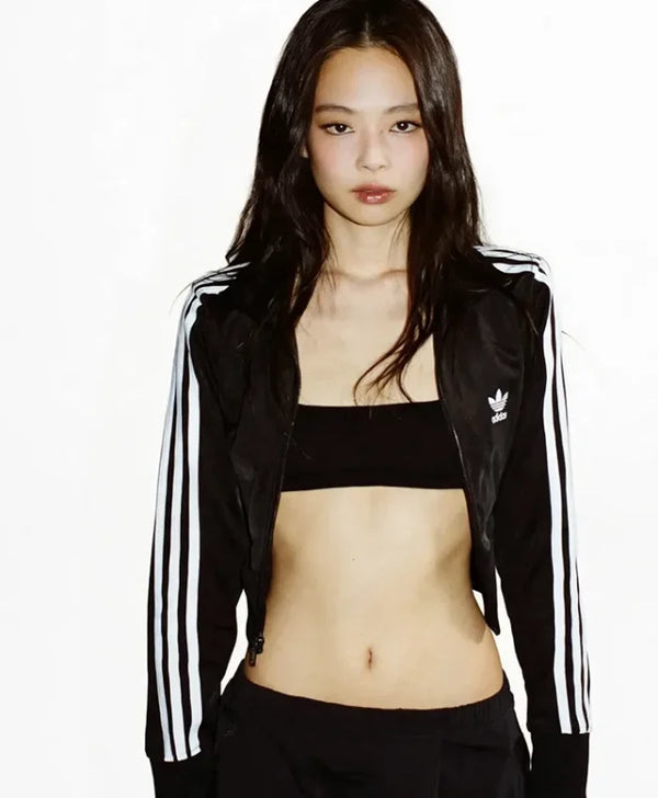 A woman modeling a black Adidas track jacket with white stripes, worn open over a black bandeau top and black pants, against a plain white background.