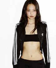 A woman modeling a black Adidas track jacket with white stripes, worn open over a black bandeau top and black pants, against a plain white background.