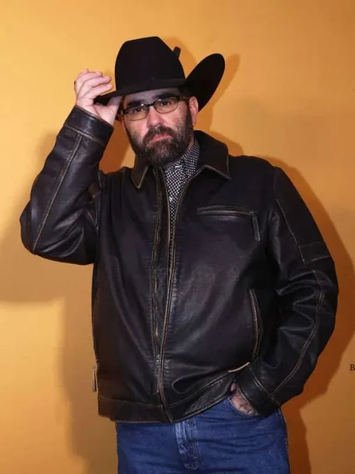 A man wearing a black leather jacket, black cowboy hat, and glasses, posing against a solid yellow background.