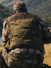 A man viewed from behind wearing a brown waxed canvas utility vest over a plaid shirt, set against a natural outdoor mountain background.
