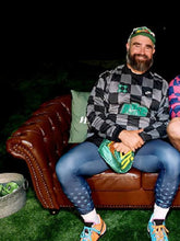 Man sitting on a brown leather couch holding a green sneaker, wearing a checkered shirt and blue jeans.