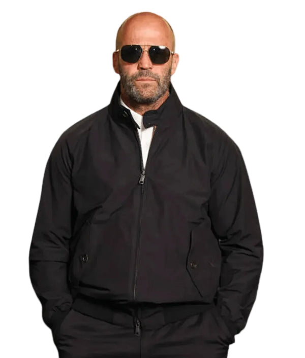 A bald man with a beard wearing black aviator sunglasses, a black zip-up jacket over a white shirt, and black pants, photographed against a plain light gray background.