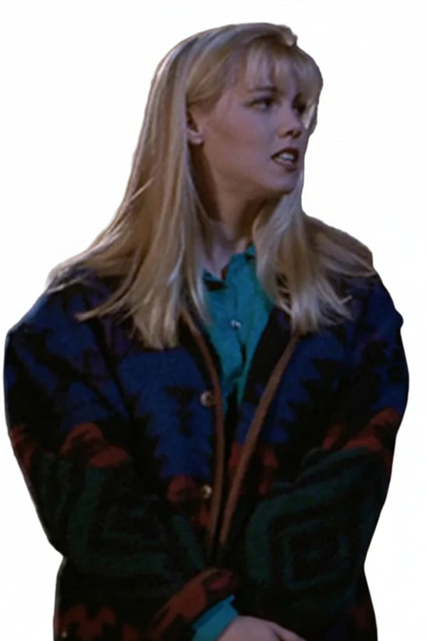 A blonde woman wearing a dark patterned cardigan over a teal shirt, standing in a dimly lit interior next to another person partially visible on the right.