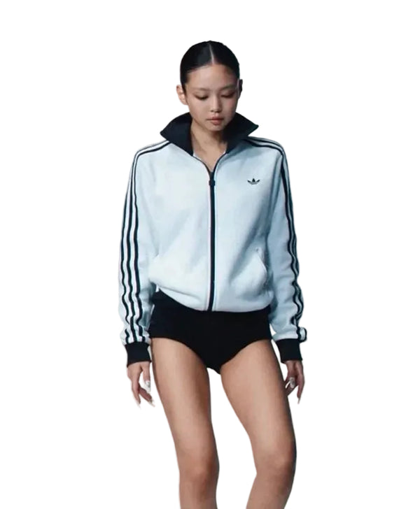 A model wearing a light blue and black Adidas track jacket with three stripes on the sleeves, paired with black shorts, against a plain white background.