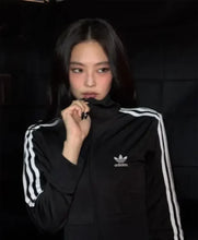 A woman wearing a black Adidas track jacket with white stripes on the sleeves and chest logo, posed against a dark background.