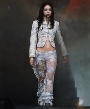 A model stands on a stage wearing a sheer, light blue and white floral lace two-piece outfit with a cropped jacket and matching ruffled pants.