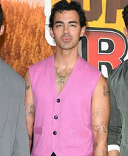 A man wearing a bright pink sleeveless knit vest over a black shirt, standing in front of a colorful event backdrop with other people partially visible at the sides.