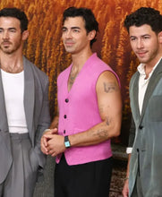 Three men standing together at what appears to be a public event or premiere, with the central figure wearing a bright pink sleeveless vest over a dark shirt and pants, flanked by two men in gray suits.