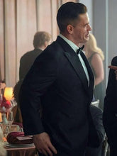 A man in a black tuxedo with a white dress shirt and black bow tie standing at a formal event, smiling and looking to the side.