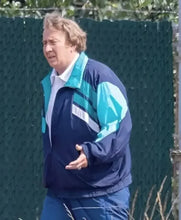 A man wearing a navy blue and turquoise athletic windbreaker jacket and matching pants, standing outdoors in front of a dark green chain-link fence.