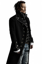 A man wearing a long black military-style coat with silver buttons and trim, layered over a dark vest and light blue jeans, set against a plain white background.