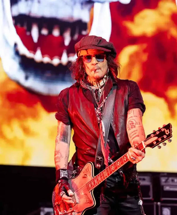A tattooed musician wearing a dark cap, sunglasses, and a dark vest holds a red electric guitar on stage in front of a large fiery graphic backdrop.