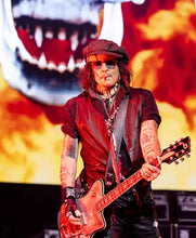 A tattooed musician wearing a dark cap, sunglasses, and a dark vest holds a red electric guitar on stage in front of a large fiery graphic backdrop.