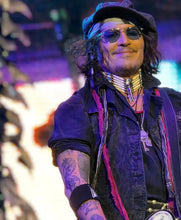 A man on stage wearing a black hat, sunglasses, layered necklaces, and a black denim vest over a dark shirt, with purple stage lighting and a blurred background.