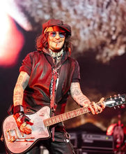 A tattooed musician on stage holding a pink electric guitar, wearing a dark cap, sunglasses, and a dark shirt with layered necklaces under dramatic red stage lighting.