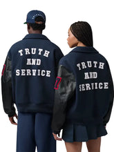 Two models shown from the back wearing matching navy blue varsity-style jackets with black leather sleeves and large white text reading 'TRUTH AND SERVICE' across the back. One model also wears a navy cap, and both are photographed against a plain light gray studio background.
