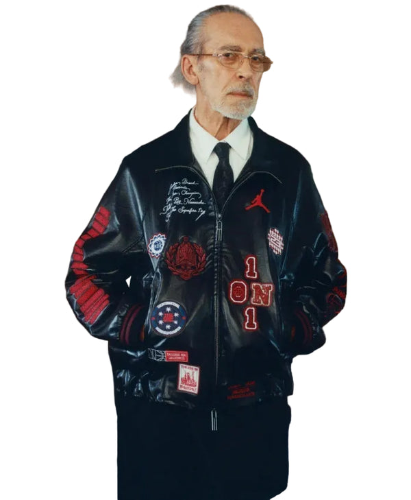 An older man with glasses, a gray beard, and a white shirt wearing a black leather bomber jacket covered in patches and red embroidery, posed against a brown textured wall.