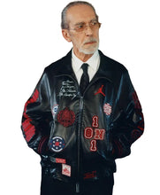 An older man with glasses, a gray beard, and a white shirt wearing a black leather bomber jacket covered in red and white patches and embroidery, including a red Jordan logo.