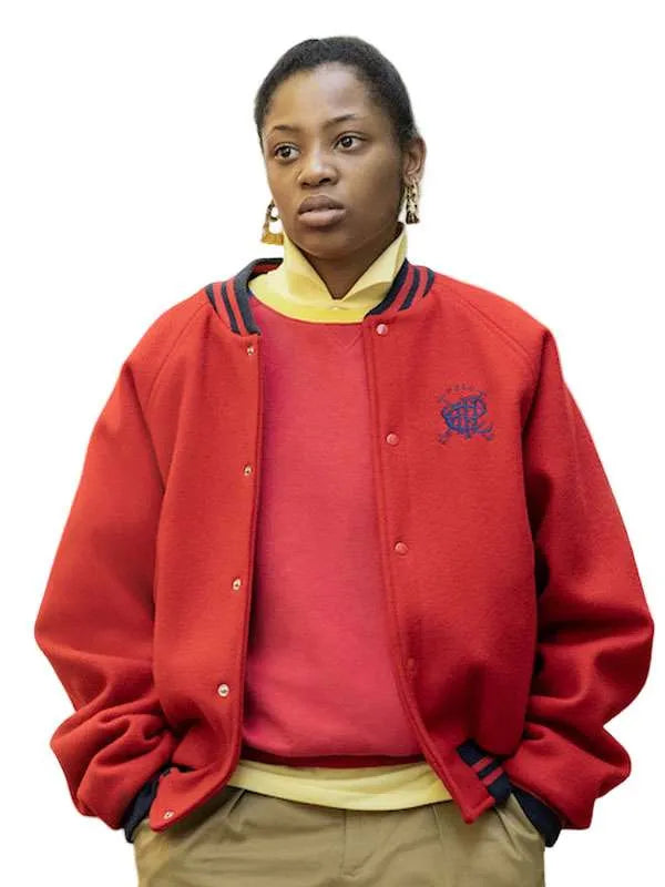 A woman wearing a red varsity-style jacket with dark navy and yellow stripes on the collar, layered over a red sweater, yellow shirt, and khaki pants, against a plain white background.