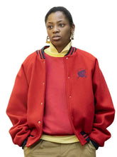 A woman wearing a red varsity-style jacket with dark navy and yellow stripes on the collar, layered over a red sweater, yellow shirt, and khaki pants, against a plain white background.