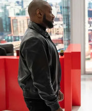 A man in a black leather jacket standing in profile in a modern office setting with a red partition wall and a city skyline visible through the windows.