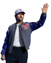 A man on a stage wearing a blue and gray varsity-style jacket with a 'Ryan' patch, a white t-shirt, dark pants, and a baseball cap, waving to a crowd while holding a microphone.