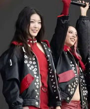 Two women on stage wearing matching black leather jackets with silver studs and red accents, over red outfits, with one holding a microphone.