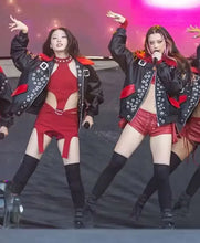 Two female dancers performing on stage wearing matching outfits: black studded leather jackets with red lining, red mini dresses or shorts, and black thigh-high socks. The background has pink and purple stage lighting.
