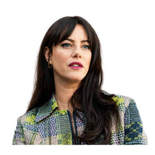 A woman with long dark brown hair and bangs, wearing a green, blue, and beige patterned tweed jacket, shown against a solid white background.